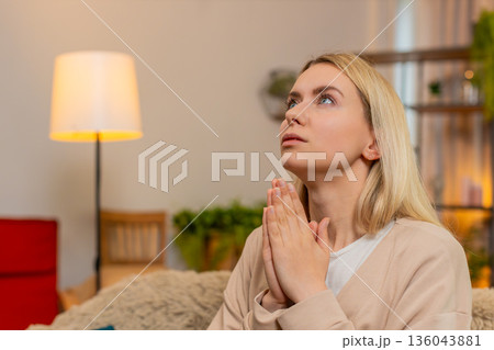 Adult woman praying at home on sofa, folding hands looking upward with sincere emotional expression 136043881