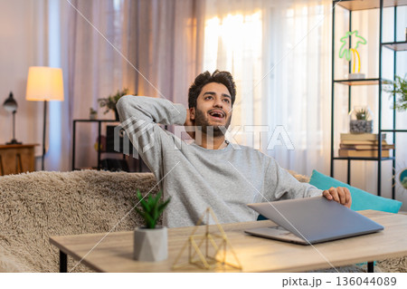 Young Indian man guy celebrating successful project completion while sitting at table with laptop 136044089