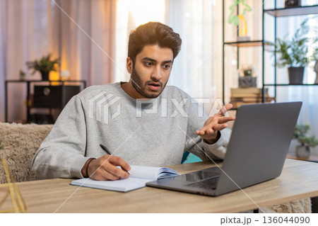 Young Indian man student attending online class through laptop while taking notes at table at home Young Indian man student attending online class through laptop while taking notes at table at home 136044090