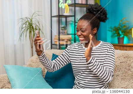 African American woman smiling waving smartphone hand gesture greeting happy indoors sofa African American woman smiling waving smartphone hand gesture greeting happy indoors sofa 136044130