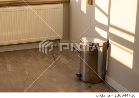 A trash can stands in the corner of a room. Sunlight enters through a window and creates shadows on the floor and wall A trash can stands in the corner of a room. Sunlight enters through a window and creates shadows on the floor and wall 136044416