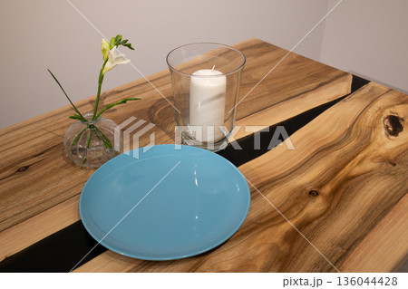 A simple table has a blue plate, a glass candle holder, and a small vase with a flower. The setting shows casual decor in a home 136044428
