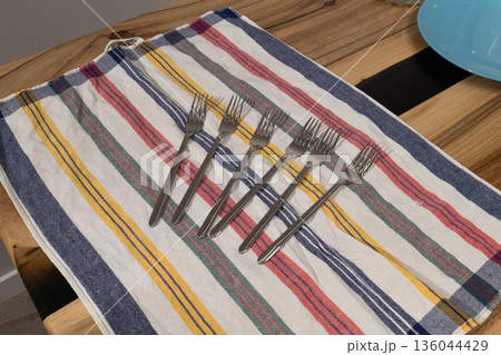 Six metal forks are arranged in a straight line on a striped dish towel in a kitchen setting. The forks are shiny and reflect light Six metal forks are arranged in a straight line on a striped dish towel in a kitchen setting. The forks are shiny and reflect light 136044429