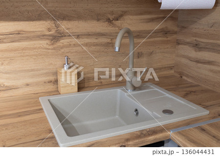 A kitchen sink sits on a wooden counter. The space shows a modern design with a soap dispenser nearby. Water faucet is ready for use A kitchen sink sits on a wooden counter. The space shows a modern design with a soap dispenser nearby. Water faucet is ready for use 136044431
