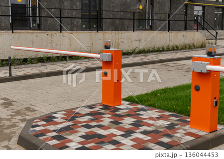 Two vibrant orange barriers stand guard at a contemporary parking lot entrance, surrounded by neatly arranged bricks and green grass Two vibrant orange barriers stand guard at a contemporary parking lot entrance, surrounded by neatly arranged bricks and green grass 136044453