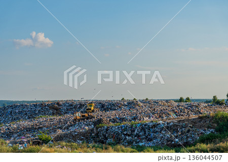 A vast landfill sprawls under a clear blue sky, piles of garbage hinting at the increasing challenge of waste management and its ecological impact A vast landfill sprawls under a clear blue sky, piles of garbage hinting at the increasing challenge of waste management and its ecological impact 136044507