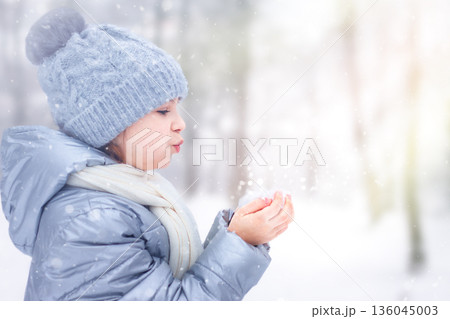 Young child in a blue winter coat blows snow from gloved hands outdoors 136045003