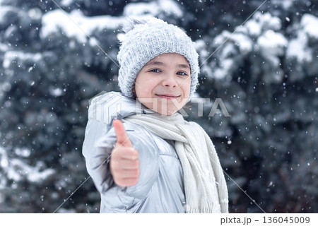 Smiling girl in winter gear gives thumbs up as snow falls in a snowy forest Smiling girl in winter gear gives thumbs up as snow falls in a snowy forest 136045009