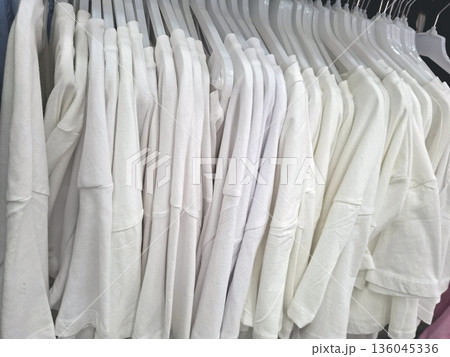 Many identical white cotton T shirts hang neatly on hangers in an eco friendly clothing store. The display shows the available options for customers in a clean manner. Many identical white cotton T shirts hang neatly on hangers in an eco friendly clothing store. The display shows the available options for customers in a clean manner. 136045336