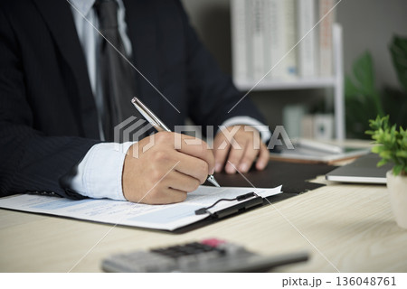 Businessman in suit signing contract document at office desk. Closeup hand holding pen for business agreement, partnership deal, legal commitment, finance investment, or insurance policy approval. 136048761
