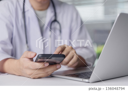 Doctor uses smartphone next to laptop in clinic office, telemedicine ehealth patient consult message schedule record management secure communication concept 136048794