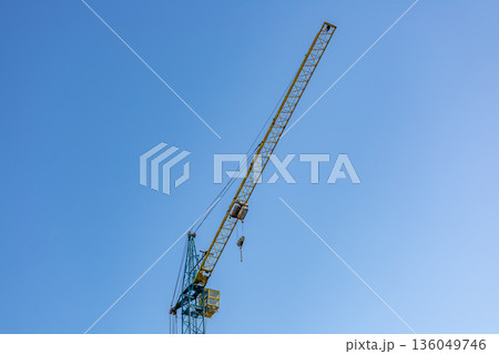 A tall construction crane towers over a bustling cityscape, reaching high into the bright blue sky on a warm sunny day 136049746