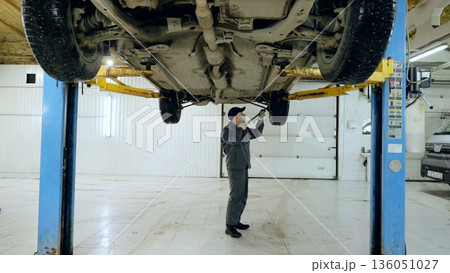 Professional mechanic checking undercarriage of lifted vehicle with crowbar, performing detailed maintenance inspection in modern auto repair workshop Professional mechanic checking undercarriage of lifted vehicle with crowbar, performing detailed maintenance inspection in modern auto repair workshop 136051027