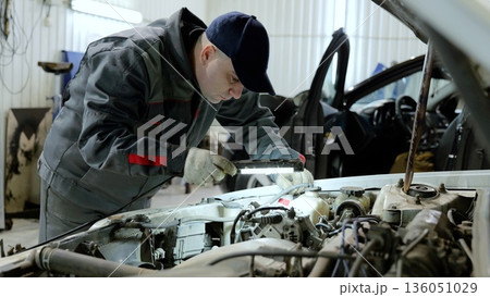 Skilled mechanic checking vehicle engine, wearing grey uniform and protective gloves, illuminating engine compartment with flashlight 136051029