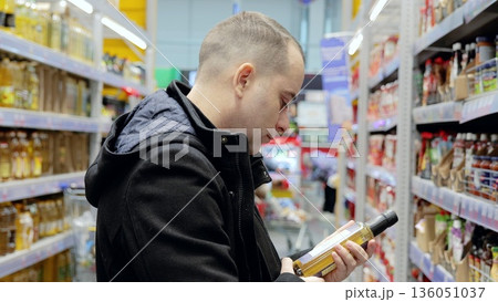Focused shopper examining olive oil bottle label, selecting quality grocery item for household needs during supermarket trip 136051037