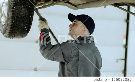 Skilled mechanic wearing protective gloves working carefully with hydraulic lever under raised vehicle, performing detailed automotive repair in workshop setting Skilled mechanic wearing protective gloves working carefully with hydraulic lever under raised vehicle, performing detailed automotive repair in workshop setting 136051080