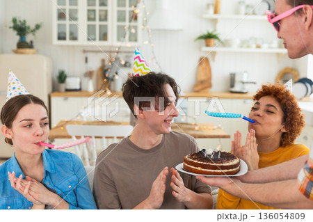 Make a wish. Man wearing party cap blowing out burning candles on birthday cake. Happy Birthday party. Group of friends wishes guy happy birthday. People celebrating birthday with party at home Make a wish. Man wearing party cap blowing out burning candles on birthday cake. Happy Birthday party. Group of friends wishes guy happy birthday. People celebrating birthday with party at home 136054809