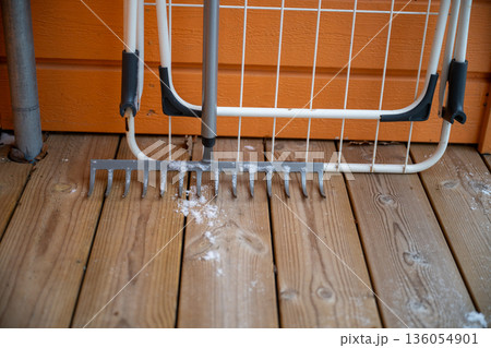 Garden rake with snow on wooden terrace against orange wall in winter Garden rake with snow on wooden terrace against orange wall in winter 136054901