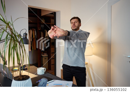 Man standing at home office desk stretching arms forward during work break 136055102