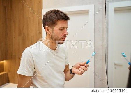 Man standing in bathroom and looking at toothbrush while facing mirror Man standing in bathroom and looking at toothbrush while facing mirror 136055104