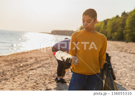 Earth day. Volunteers activists team collects garbage cleaning of beach coastal zone. Woman puts plastic bottle trash in garbage bag on ocean shore. Environmental conservation coastal zone cleaning 136055310
