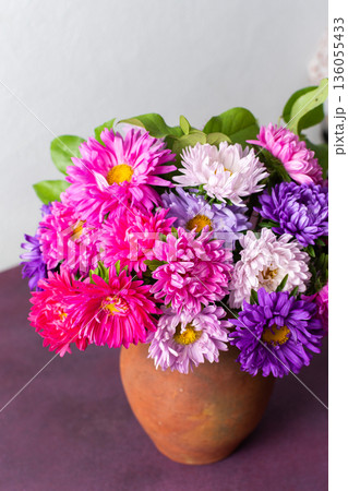 A clay pot holds a mix of colorful flowers arranged neatly on a pink table. The setting is bright with a plain background, showing natural beauty and simplicity 136055433