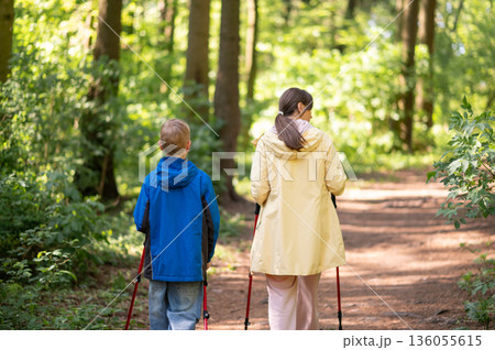 Rear view of mother and 11-year-old son hiking with Nordic trekking poles through a sunny forest trail Rear view of mother and 11-year-old son hiking with Nordic trekking poles through a sunny forest trail 136055615