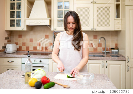 Smiling woman cutting cucumber for fresh vegetable salad in cozy kitchen. Healthy lifestyle, home cooking, diet and proper nutrition 136055783