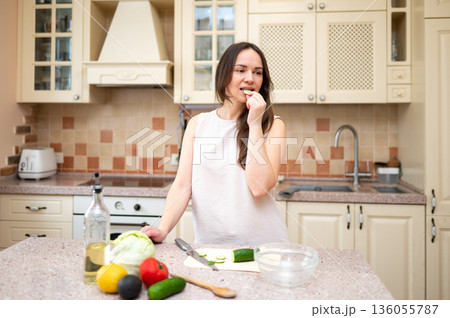 Young woman in bright kitchen eating cucumber and preparing salad. Fresh vegetables and ingredients on counter suggest healthy and balanced diet 136055787
