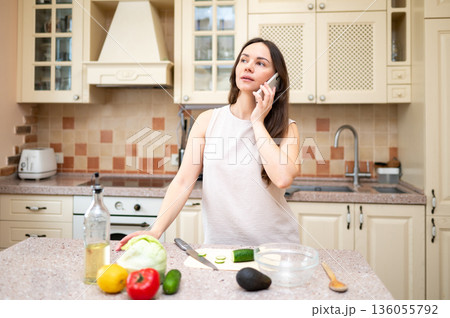 young woman in bright kitchen talking on phone and smiling, planning to cook salad. Fresh vegetables and board are on table 136055792