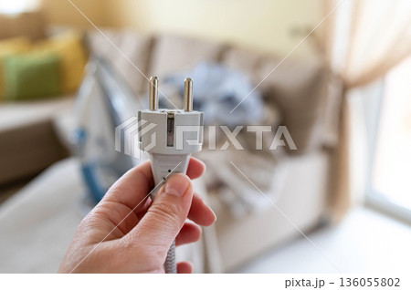 A plug with a power cord from a disconnected iron in a woman's hand against the background of an ironing board with an iron and a pile of crumpled laundry A plug with a power cord from a disconnected iron in a woman's hand against the background of an ironing board with an iron and a pile of crumpled laundry 136055802