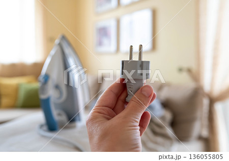 A plug with a power cord from a disconnected iron in a woman's hand against the background of an ironing board with an iron and a pile of crumpled laundry 136055805