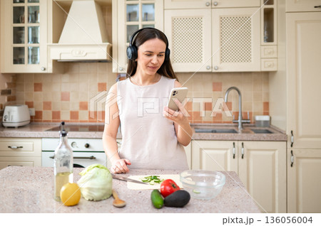 Woman using smartphone while preparing vegetables in kitchen. Wearing headphones, she appears focused. Fresh ingredients like lettuce, avocado, and tomato on the counter. Woman using smartphone while preparing vegetables in kitchen. Wearing headphones, she appears focused. Fresh ingredients like lettuce, avocado, and tomato on the counter. 136056004