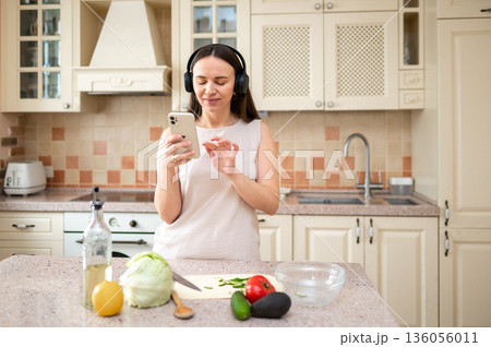 Woman using smartphone while preparing vegetables in kitchen. Wearing headphones, she appears focused. Fresh ingredients like lettuce, avocado, and tomato on the counter. Woman using smartphone while preparing vegetables in kitchen. Wearing headphones, she appears focused. Fresh ingredients like lettuce, avocado, and tomato on the counter. 136056011