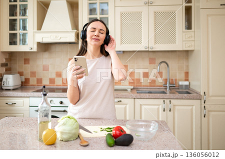 Woman listens to music with headphones, smiling while holding smartphone in kitchen. Fresh vegetables and oil bottle on counter. Relaxed and joyful mood during cooking. Woman listens to music with headphones, smiling while holding smartphone in kitchen. Fresh vegetables and oil bottle on counter. Relaxed and joyful mood during cooking. 136056012
