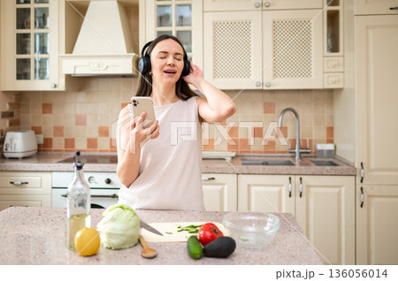 Woman listens to music with headphones, smiling while holding smartphone in kitchen. Fresh vegetables and oil bottle on counter. Relaxed and joyful mood during cooking. 136056014