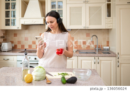 Woman holding a tomato and looking at her phone in the kitchen. She wears headphones, multitasking between cooking and technology, showing a relaxed and focused expression. Woman holding a tomato and looking at her phone in the kitchen. She wears headphones, multitasking between cooking and technology, showing a relaxed and focused expression. 136056024