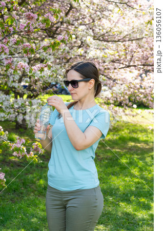 Young woman wearing sunglasses drinking water from bottle outdoors in blooming spring park Young woman wearing sunglasses drinking water from bottle outdoors in blooming spring park 136056107