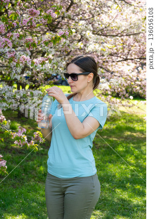 Young woman wearing sunglasses drinking water from bottle outdoors in blooming spring park Young woman wearing sunglasses drinking water from bottle outdoors in blooming spring park 136056108