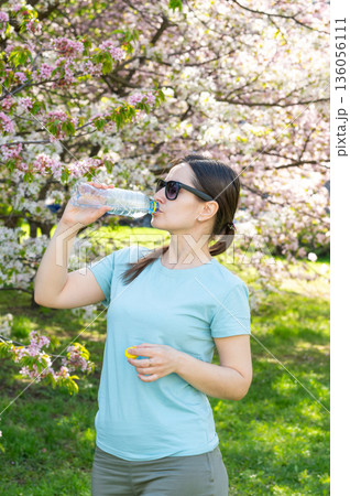 Young woman wearing sunglasses drinking water from bottle outdoors in blooming spring park Young woman wearing sunglasses drinking water from bottle outdoors in blooming spring park 136056111