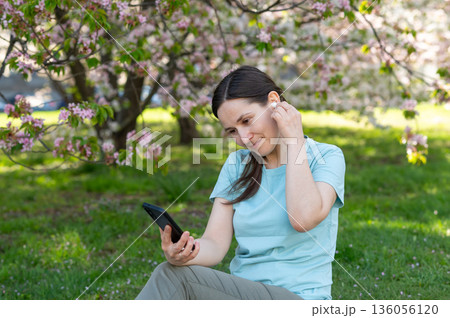 Young woman sitting on blanket in blooming spring park and using wireless headphones and smartphone listening to music Young woman sitting on blanket in blooming spring park and using wireless headphones and smartphone listening to music 136056120
