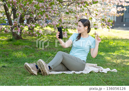 Young woman sits on a blanket in a blooming spring park, talking on a video call via smartphone 136056126