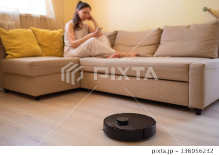 A robot vacuum cleaner washes the floor while a young woman uses her smartphone while sitting on a cozy beige sofa. Smart home, automation and modern technologies in everyday life A robot vacuum cleaner washes the floor while a young woman uses her smartphone while sitting on a cozy beige sofa. Smart home, automation and modern technologies in everyday life 136056232