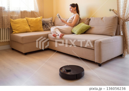 A robot vacuum cleaner washes the floor while a young woman reads a book sitting on the sofa. Smart home, automation and modern technologies in everyday life 136056236