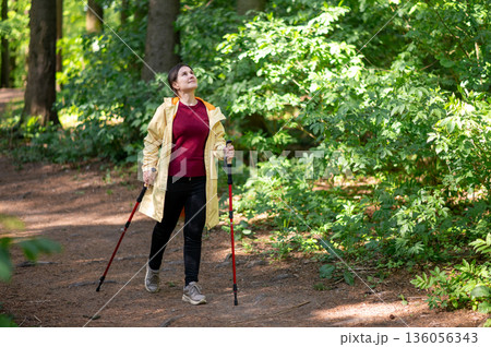 Joyful woman practicing Nordic walking with poles on a sunlit forest trail, combining fitness and nature for a healthy lifestyle 136056343