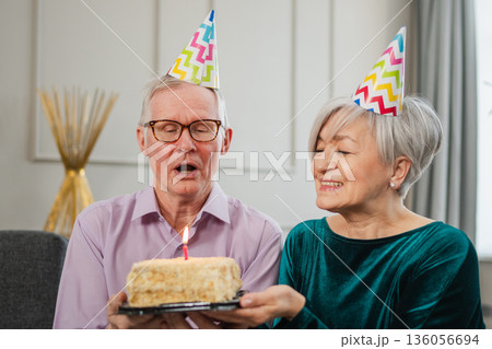 Make a wish. Family senior couple in party hat cap celebrating birthday anniversary together at home. Old man blowing out burning candles on birthday cake. Old woman wishes husband happy birthday 136056694