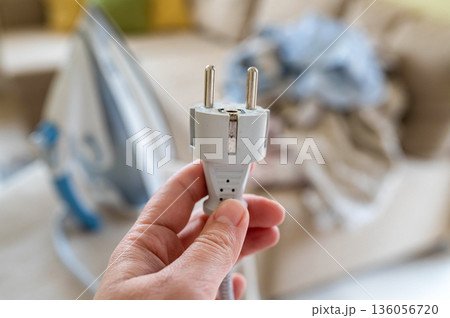 A plug with a power cord from a disconnected iron in a woman's hand against the background of an ironing board with an iron and a pile of crumpled laundry A plug with a power cord from a disconnected iron in a woman's hand against the background of an ironing board with an iron and a pile of crumpled laundry 136056720