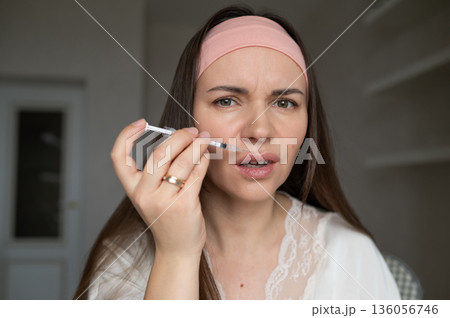 A young woman in a silk robe and headband applies a beauty injection to her face while looking in the mirror. Home skincare and aesthetic procedures A young woman in a silk robe and headband applies a beauty injection to her face while looking in the mirror. Home skincare and aesthetic procedures 136056746