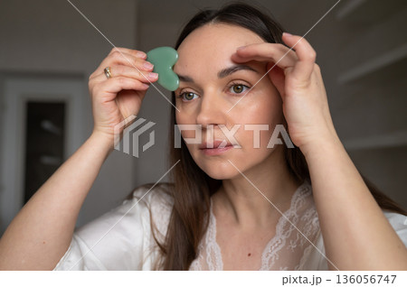 Young woman in silk robe doing facial massage with gua sha scraper while looking in mirror. Home skin care and aesthetic procedures Young woman in silk robe doing facial massage with gua sha scraper while looking in mirror. Home skin care and aesthetic procedures 136056747