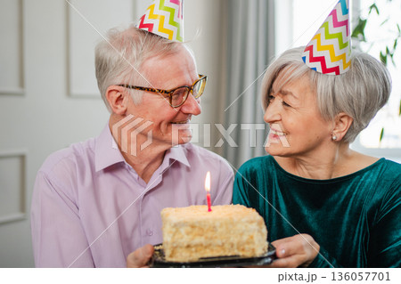 Make a wish. Family senior couple in party hat cap celebrating birthday anniversary together at home. Old woman blowing out burning candles on birthday cake. Old man wishes wife happy birthday Make a wish. Family senior couple in party hat cap celebrating birthday anniversary together at home. Old woman blowing out burning candles on birthday cake. Old man wishes wife happy birthday 136057701